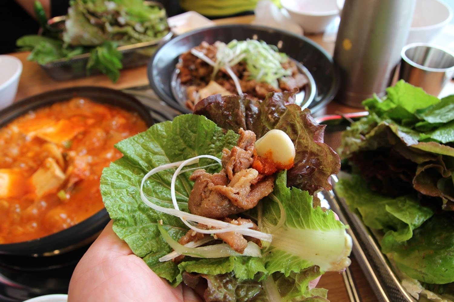 韩国传统生菜包饭，采用生菜包裹猪肉、卷心菜、泡菜和大蒜，搭配豆腐汤和桌上多种配菜，展示正宗的韩国饮食文化。