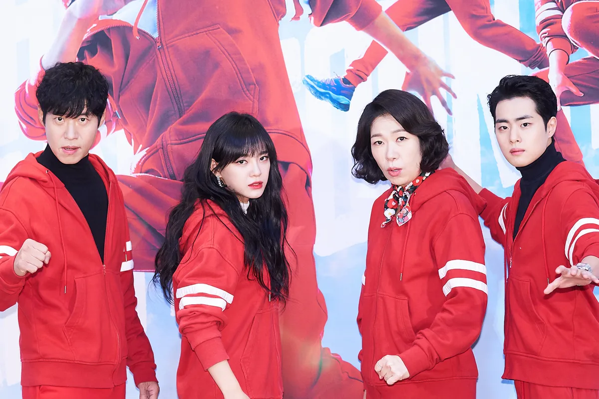 四名演員穿著紅色運動服,站在《驅魔麵館》的海報前。