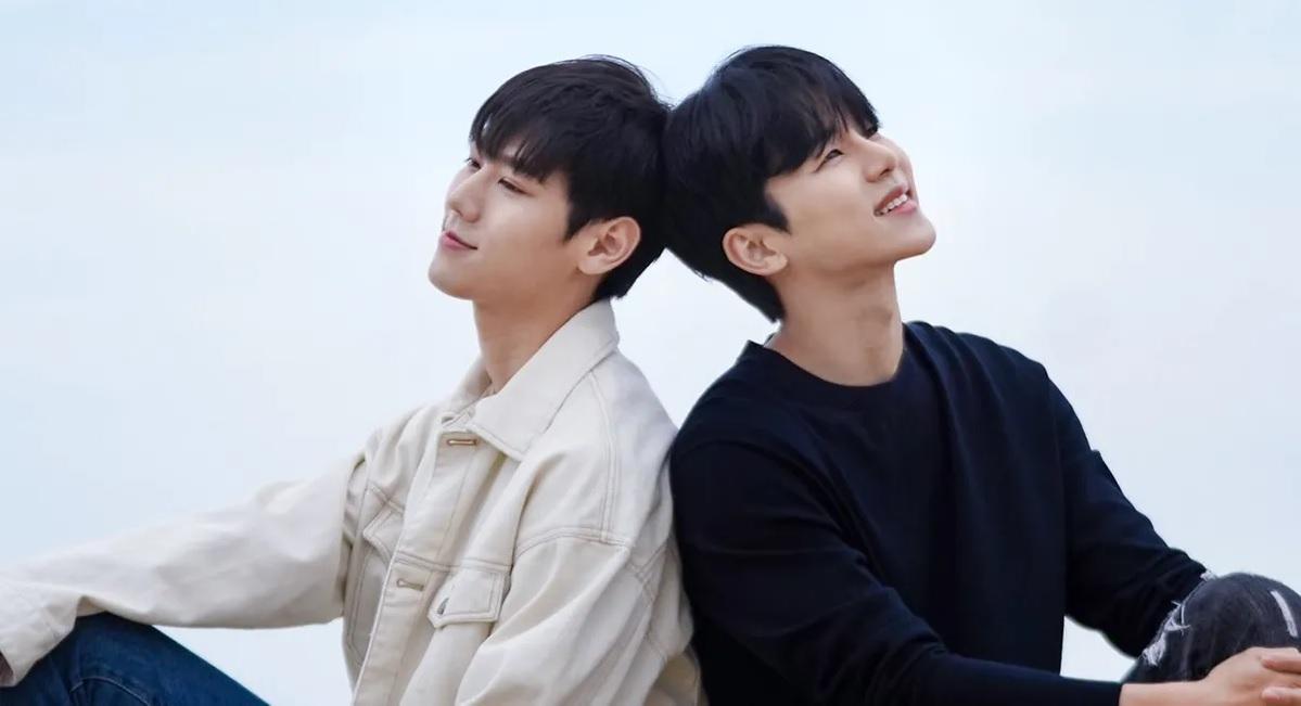 ชายสองคนในชุดลำลองใส่เสื้อผ้าสีขาวและสีดำ นั่งพิงกันบนพื้นที่มีวิวด้านหลังเป็นท้องฟ้า ทำให้ดูผ่อนคลายและเรียบง่าย