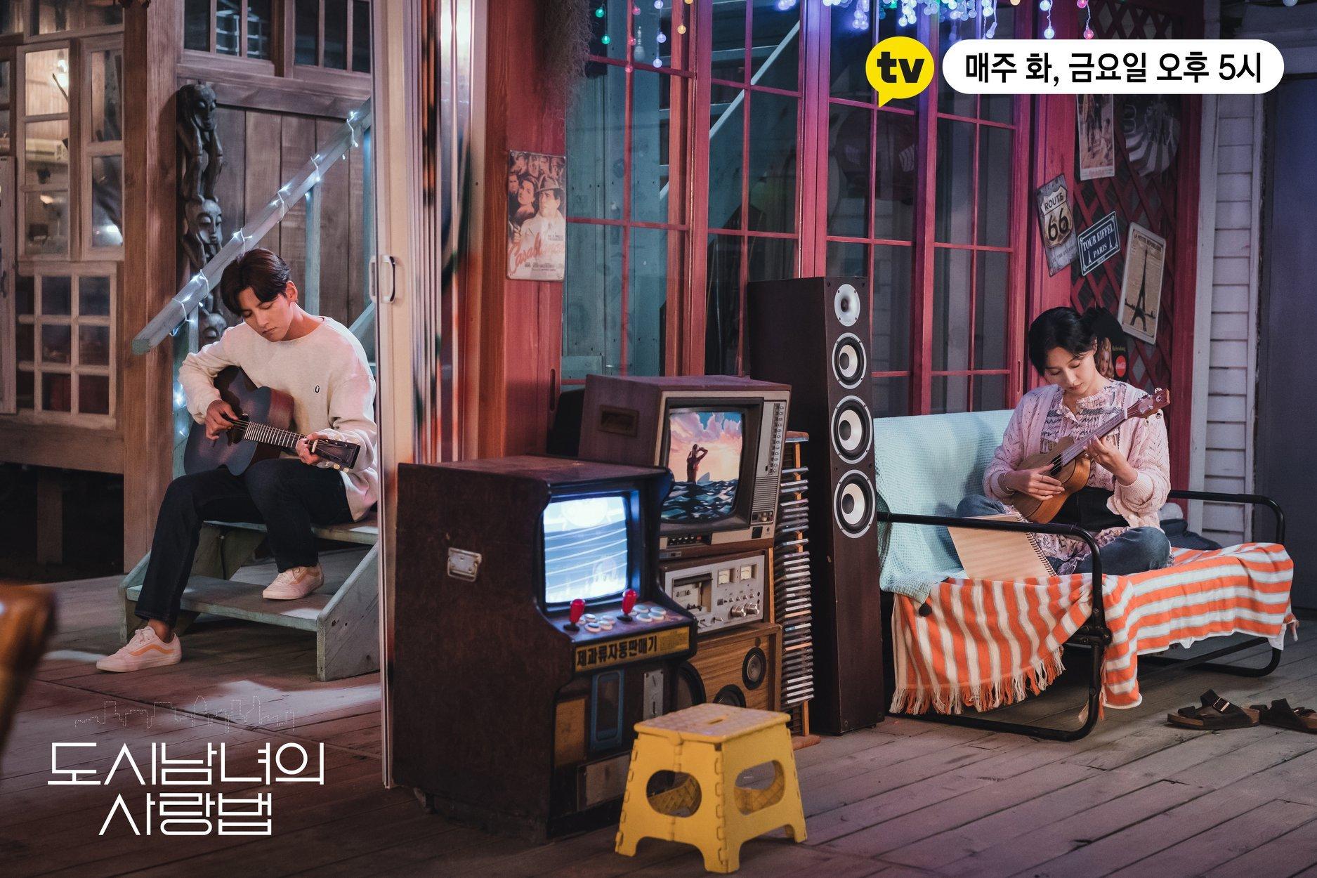 คนสองคนกำลังนั่งในบรรยากาศแบบวินเทจ โดยมีคนหนึ่งเล่นกีตาร์และอีกคนหนึ่งเล่นอูคูเลเล่อยู่บนเก้าอี้ใต้แสงไฟประดับ