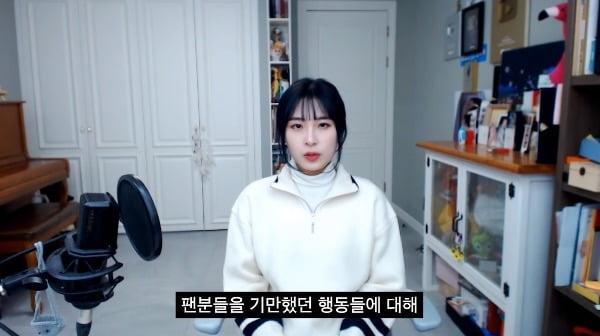 YouTubeの映像の場面。椅子に座り、白いセーターを着た人物が、カメラに向かって韓国語の字幕を持つ映像