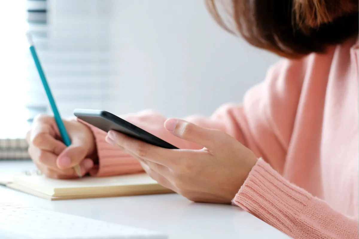 มือของผู้หญิงกำลังถือสมาร์ทโฟนขณะที่ใช้ดินสอเขียนบนสมุดบันทึก เสื้อแขนยาวสีชมพูแสดงให้เห็นสภาพอากาศหนาวเย็น