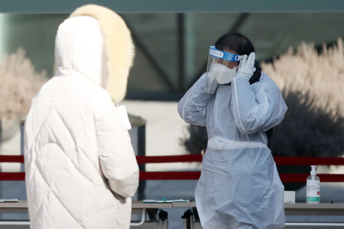 防疫人員身穿防護衣，面部帶有透明面罩，與另一位穿著白色衣物的市民對話，場景中有洗手液顯示重視衞生的措施。