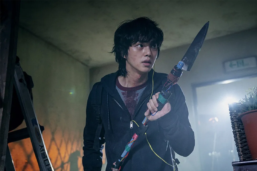 韓國電視劇《Sweet Home》中一位角色在走廊中持武器站立,背景中可見模糊的逃生出口標誌和梯子,營造出緊張的戲劇場景,來自Netflix的圖片。