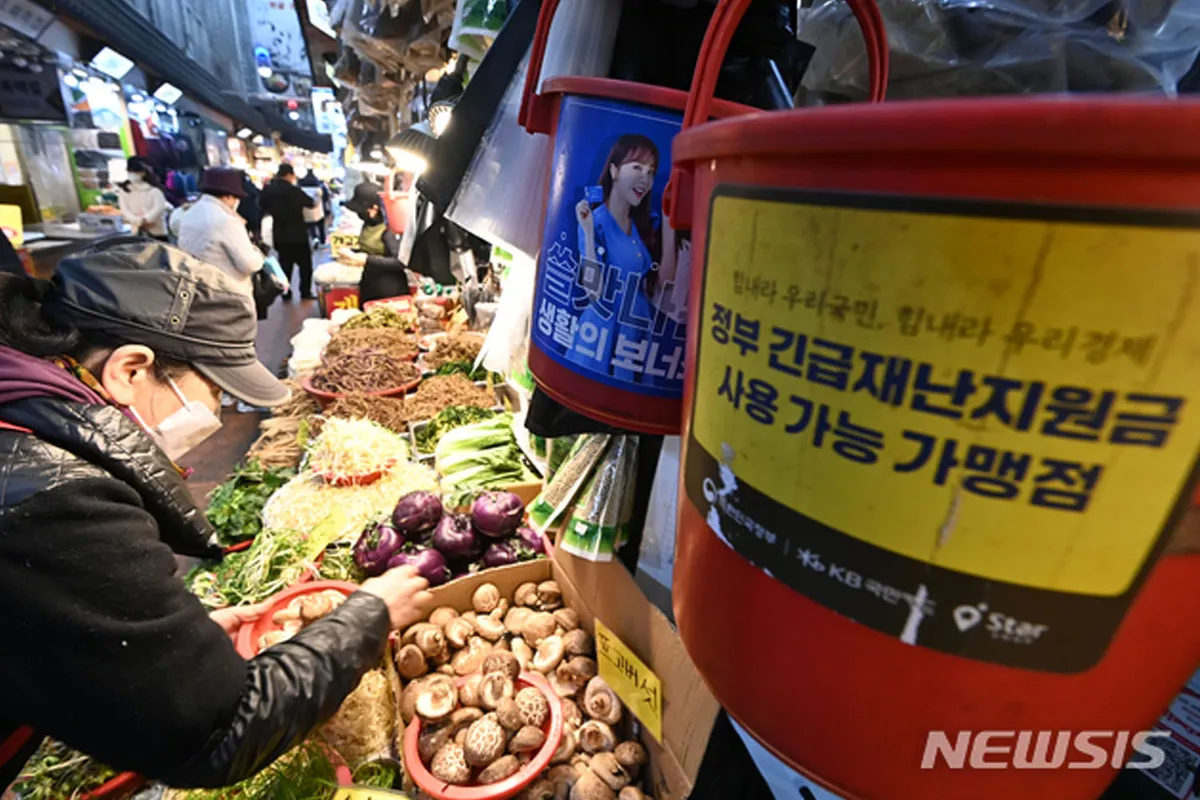 ตลาดที่ผู้คนกำลังเลือกซื้อของพร้อมด้วยถังบริจาคที่เกี่ยวข้องกับโครงการเงินช่วยเหลือ บ่งบอกถึงสถานการณ์ทางเศรษฐกิจหรือการสนับสนุนจากรัฐบาลในเกาหลี
