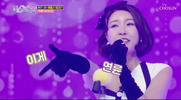 ミストロット2のステージ上で紫色の背景をバックに黒い手袋とヘッドアクセサリーを着用した女性がマイクを持ってポーズを取っている様子。