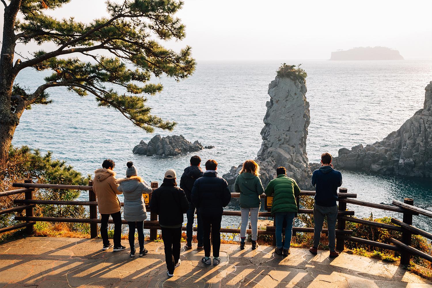 ผู้คนยืนชมวิวมหาสมุทรบนเกาะเชจูที่มีแท่นหินสูงเด่นเป็นเอกลักษณ์