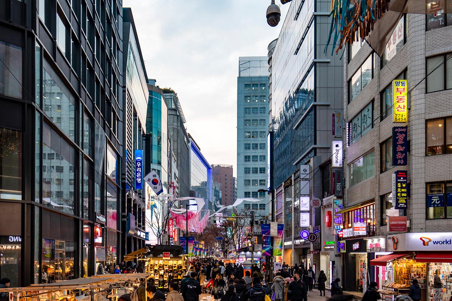 A bustling street in Incheon filled with people, tall buildings on either side adorned with colorful signs and advertisements.