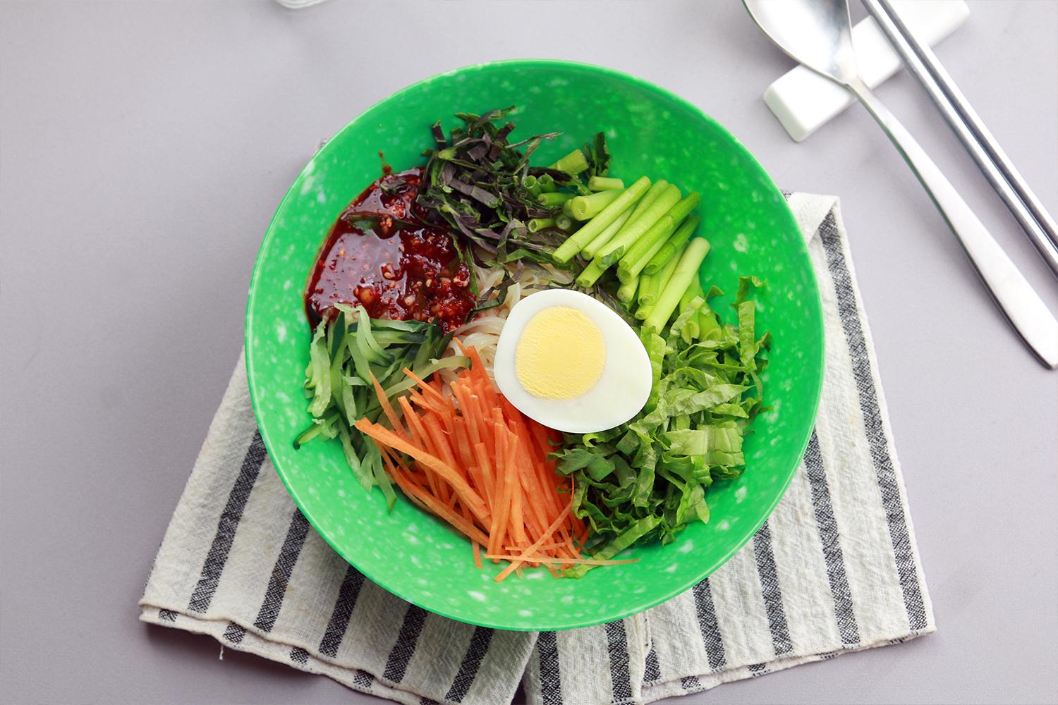A vibrant bowl of Jjolmyeon, spicy cold noodles with colorful vegetables and a boiled egg in Incheon.