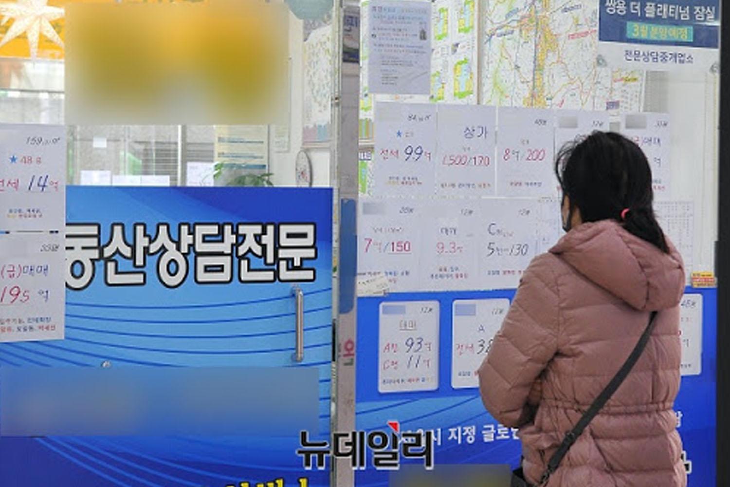 ผู้หญิงยืนหน้าร้านนายหน้าที่แสดงราคาซื้อขายอสังหาริมทรัพย์ในเกาหลีใต้