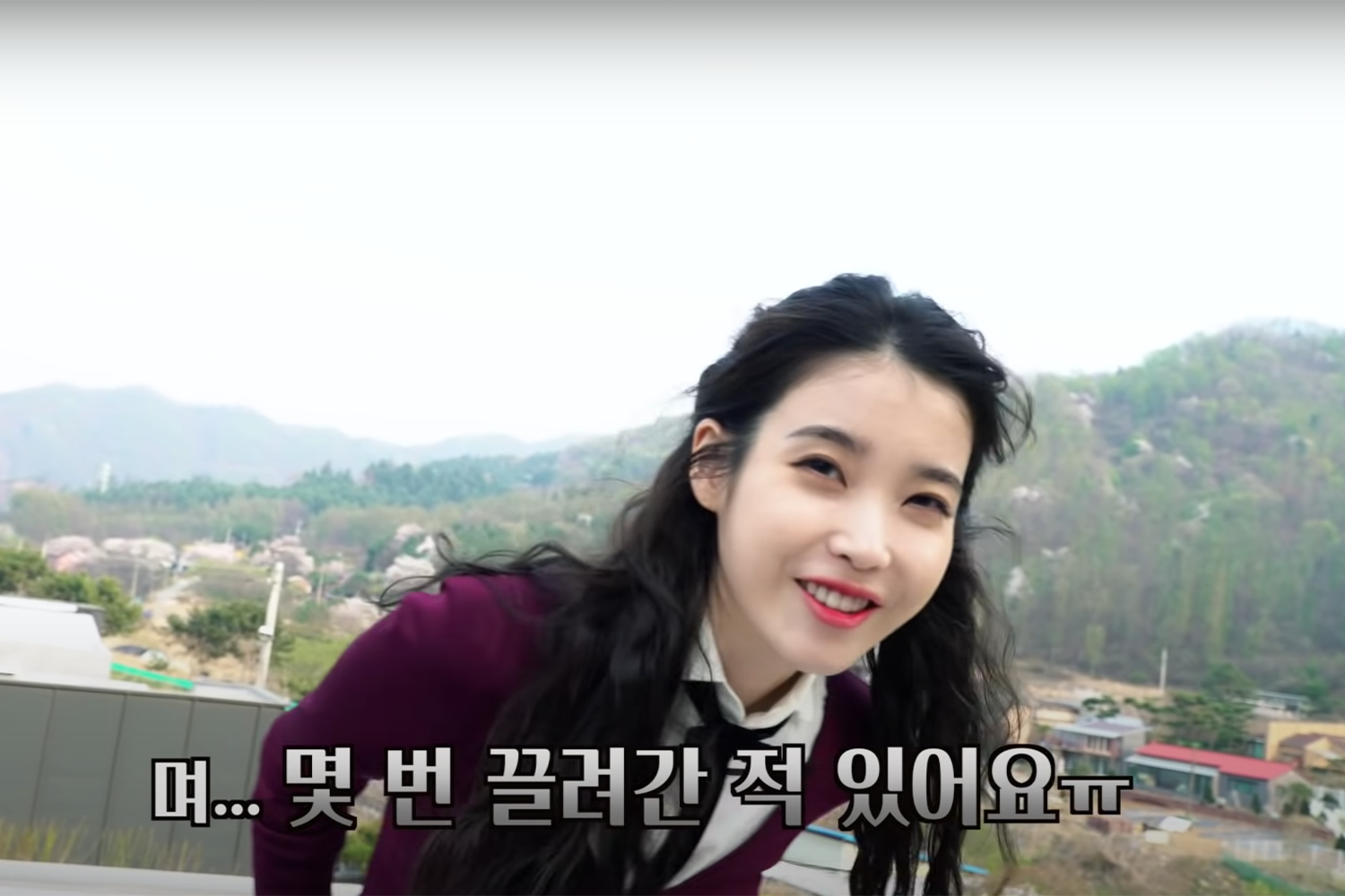 IU身穿校服在户外環境中,背景為山景和市區景觀。韓文字幕表示她過去曾經被帶走數次。