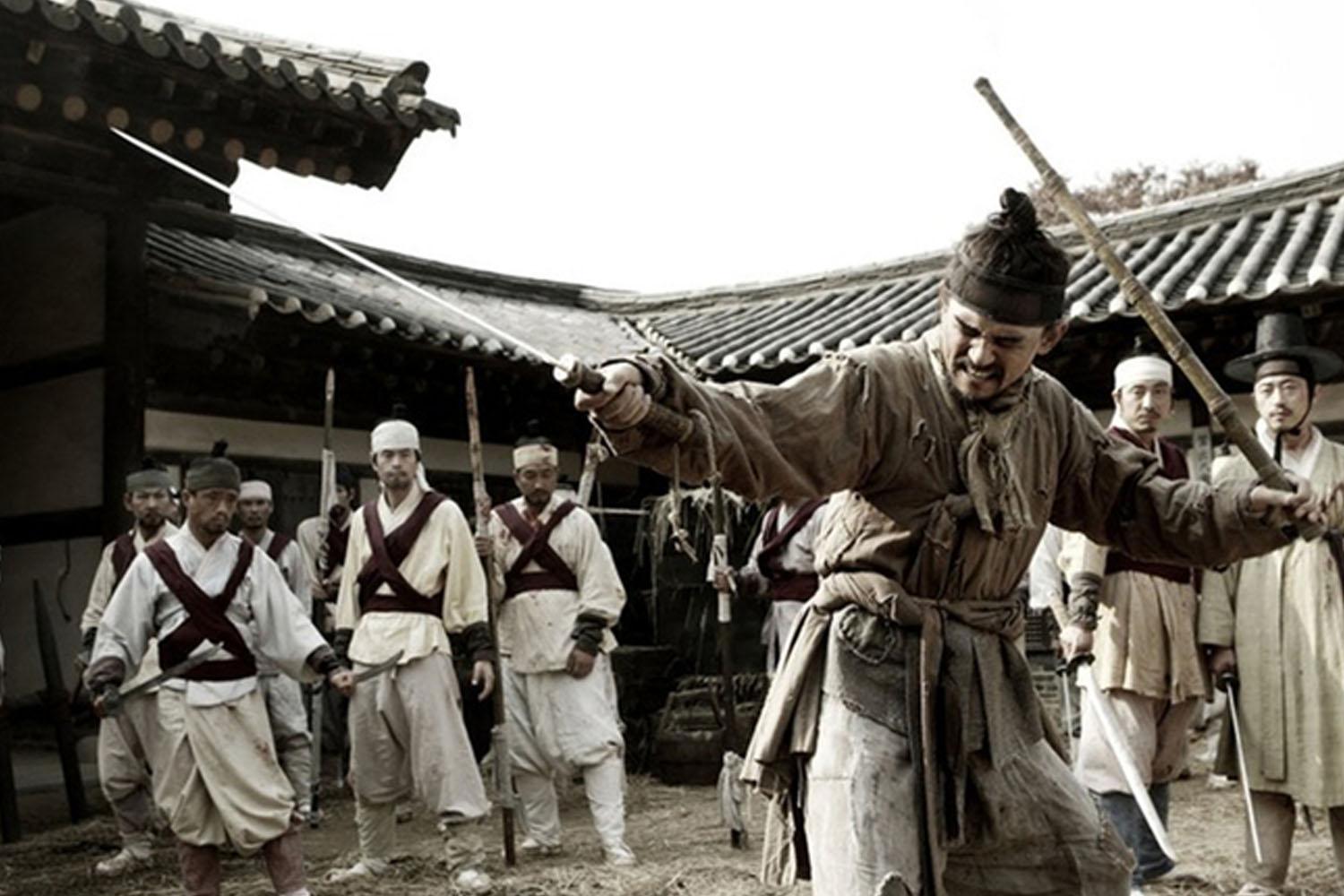 กลุ่มนักรบสวมชุดดั้งเดิมในยุคโชซอนถืออาวุธยืนอยู่ในลานของอาคารสไตล์เกาหลีแบบดั้งเดิม แสดงถึงบรรยากาศยุคเก่าและการต่อสู้ในประวัติศาสตร์เกาหลี