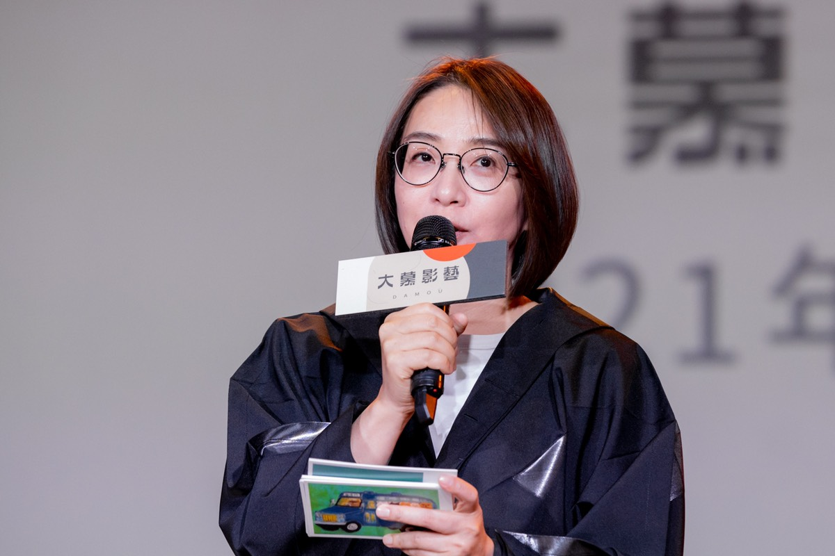 大慕影藝的一位成員手持麥克風在年度發表會上演講的場景，背後是發表會的背景板。