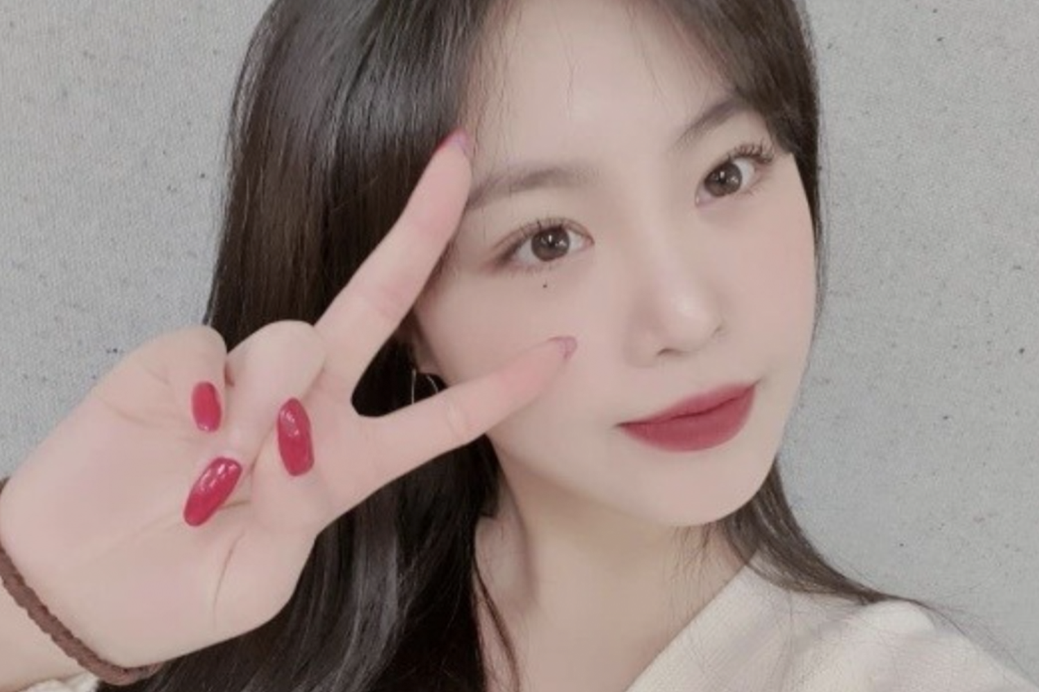 (G)I-DLE 成員穗珍用紅色指甲擺出剪刀手手勢，穿著米色上衣，背景為灰色牆壁。