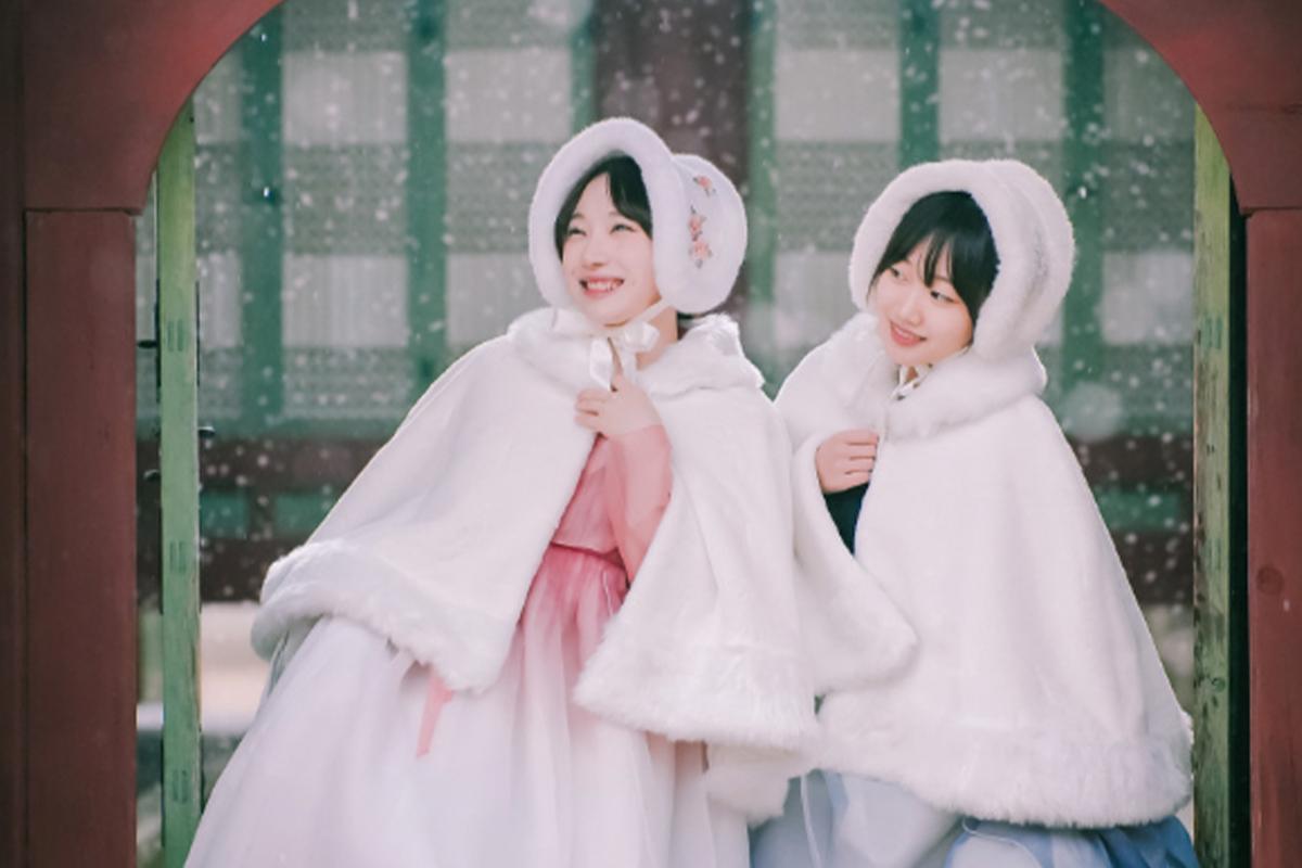 Two women in traditional hanbok, wearing white cloaks, standing under a wooden archway with snowfall around them.