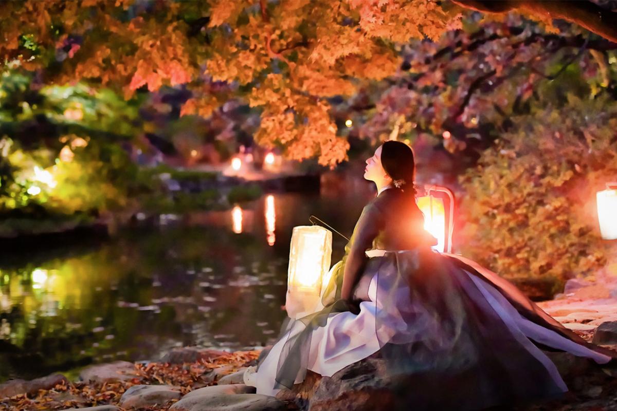 Woman in a hanbok sitting near a pond under colorful autumn leaves, with warm lantern lights illuminating the area.