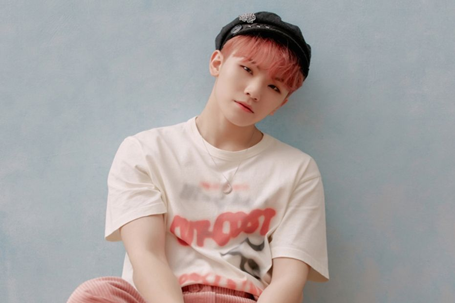 SEVENTEEN 成員 WOOZI 坐在灰藍色背景前，穿著印有紅色字樣的白色T恤及粉紅色長褲。