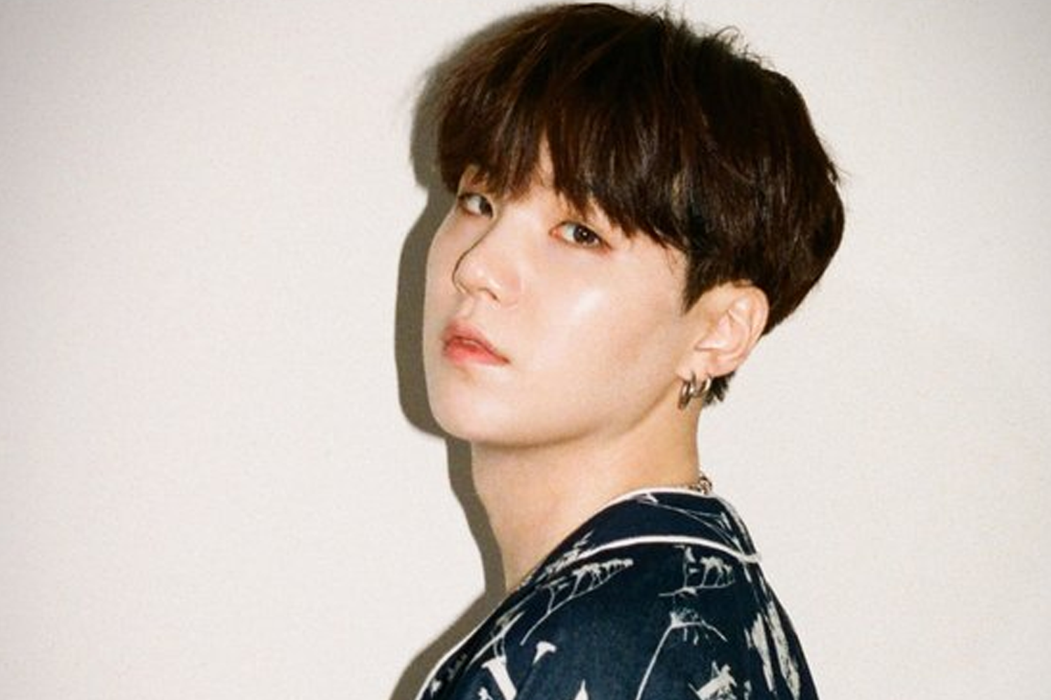 BTS 成員 SUGA 穿著印花上衣，背景為淺色牆壁。