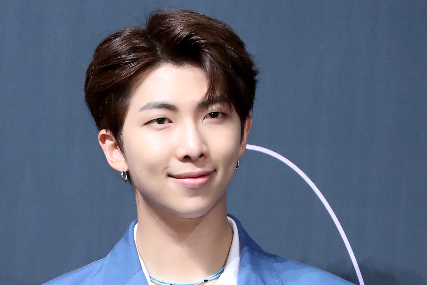 BTS 成員 RM 在舞台背景前，穿著藍色西裝，背景為深色。