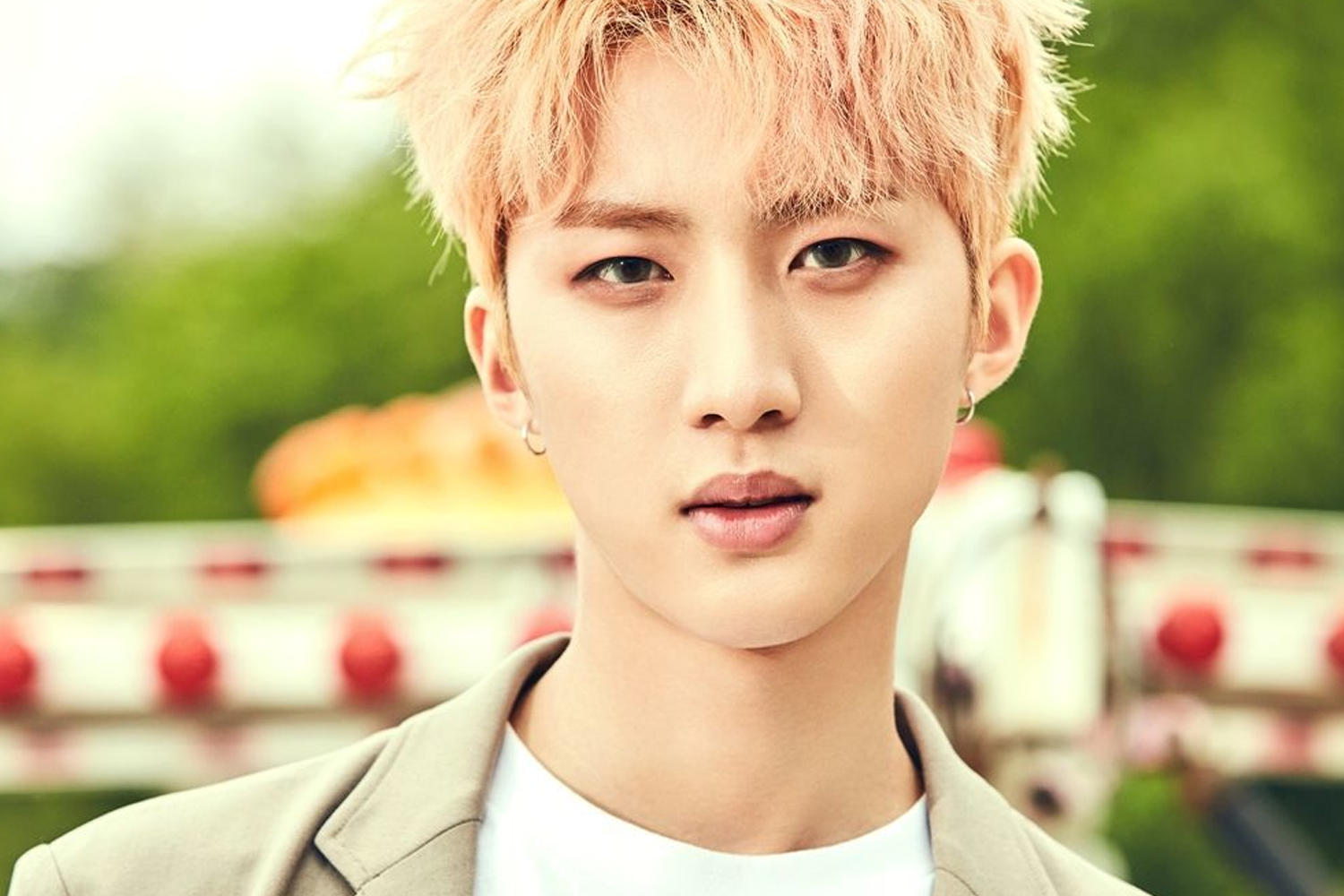 Pentagon 成員 Hui 站在室外遊樂園背景前，身穿米色西裝，頭髮是金色。