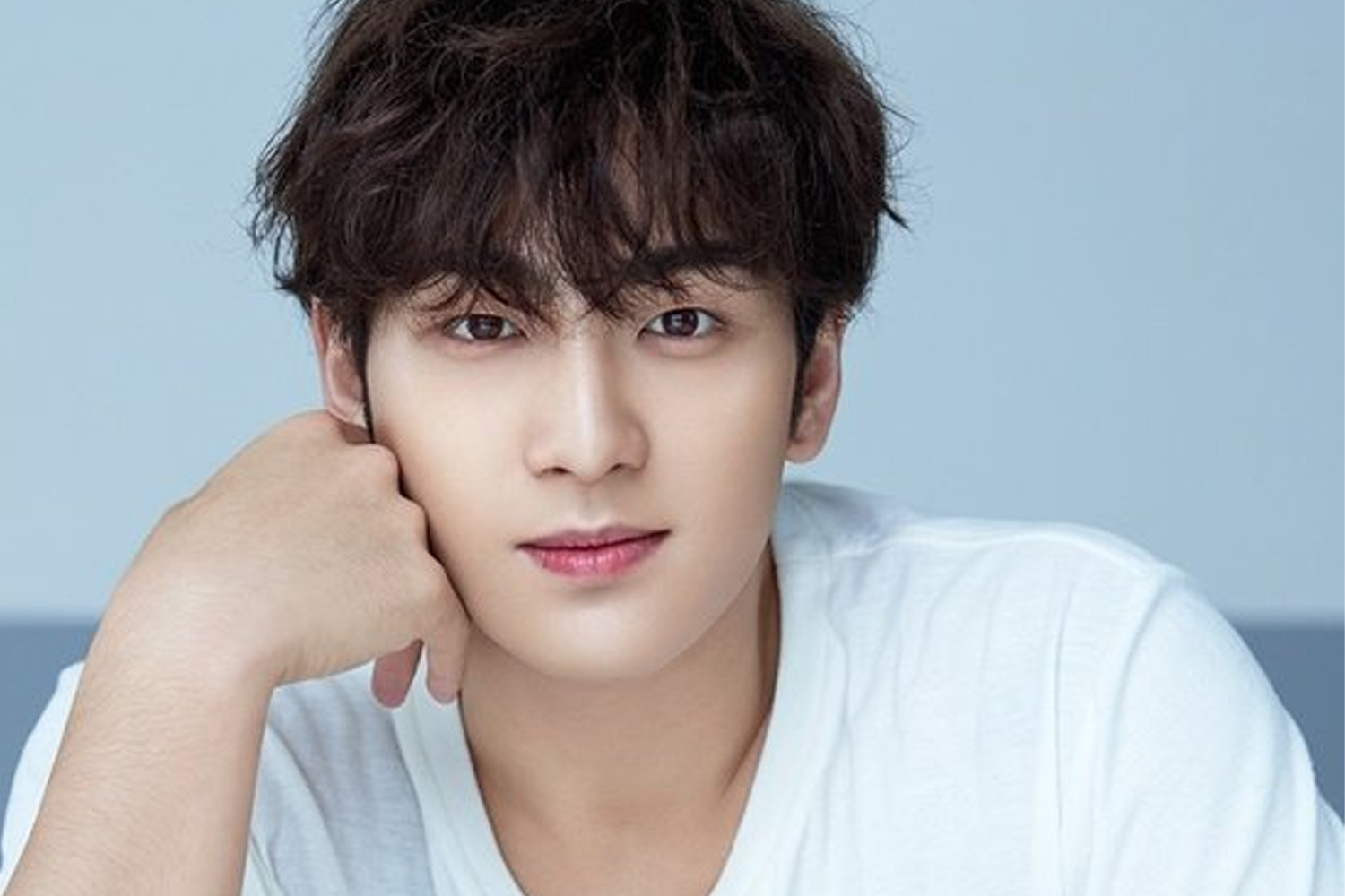 NU'EST 成員白虎坐在藍色背景前，穿著白色T恤，手撐着臉。