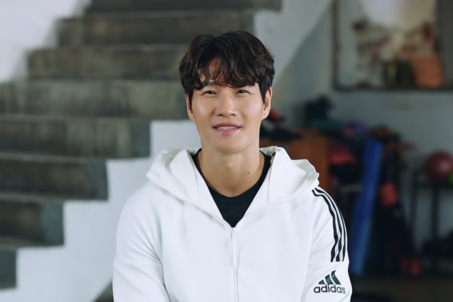 金鍾國穿著白色運動服坐在樓梯前，背景是運動器材。