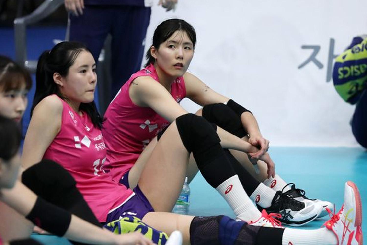 นักกีฬาวอลเลย์บอลนั่งพักข้างสนามในชุดสีชมพูขณะสวมรองเท้ากีฬาและสนับหน้าแข้ง