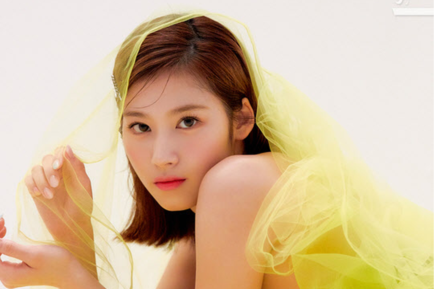 TWICE成員Sana身著亮黃色的紗質上衣，手持透明面紗，展現出她甜美和可愛的一面。