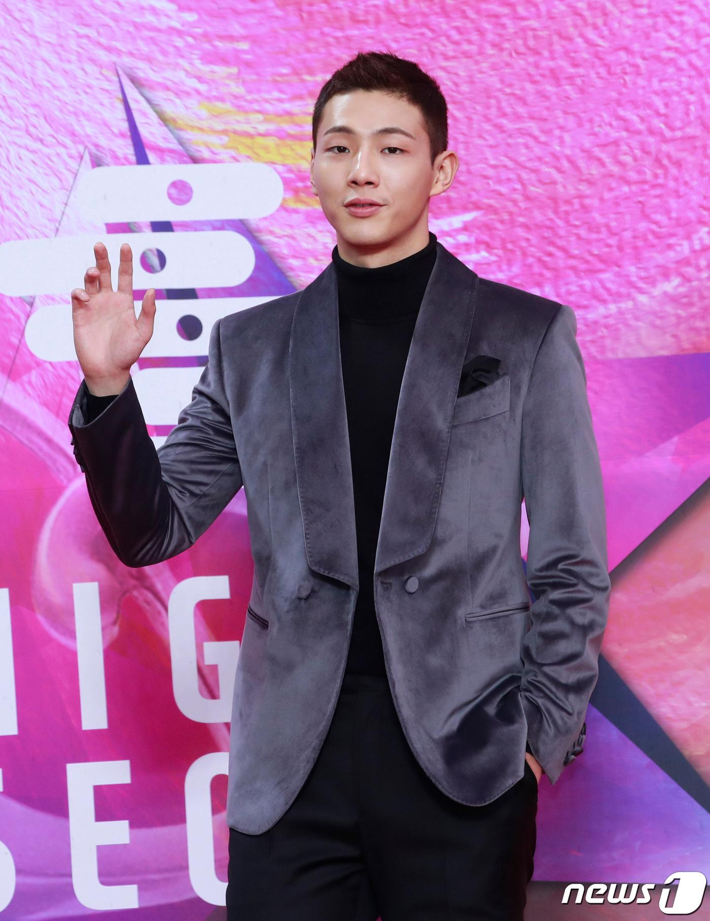 グレーのベルベットジャケットを着た男性が、背景にピンクのイベントのバナーがある場所で片手を上げている。