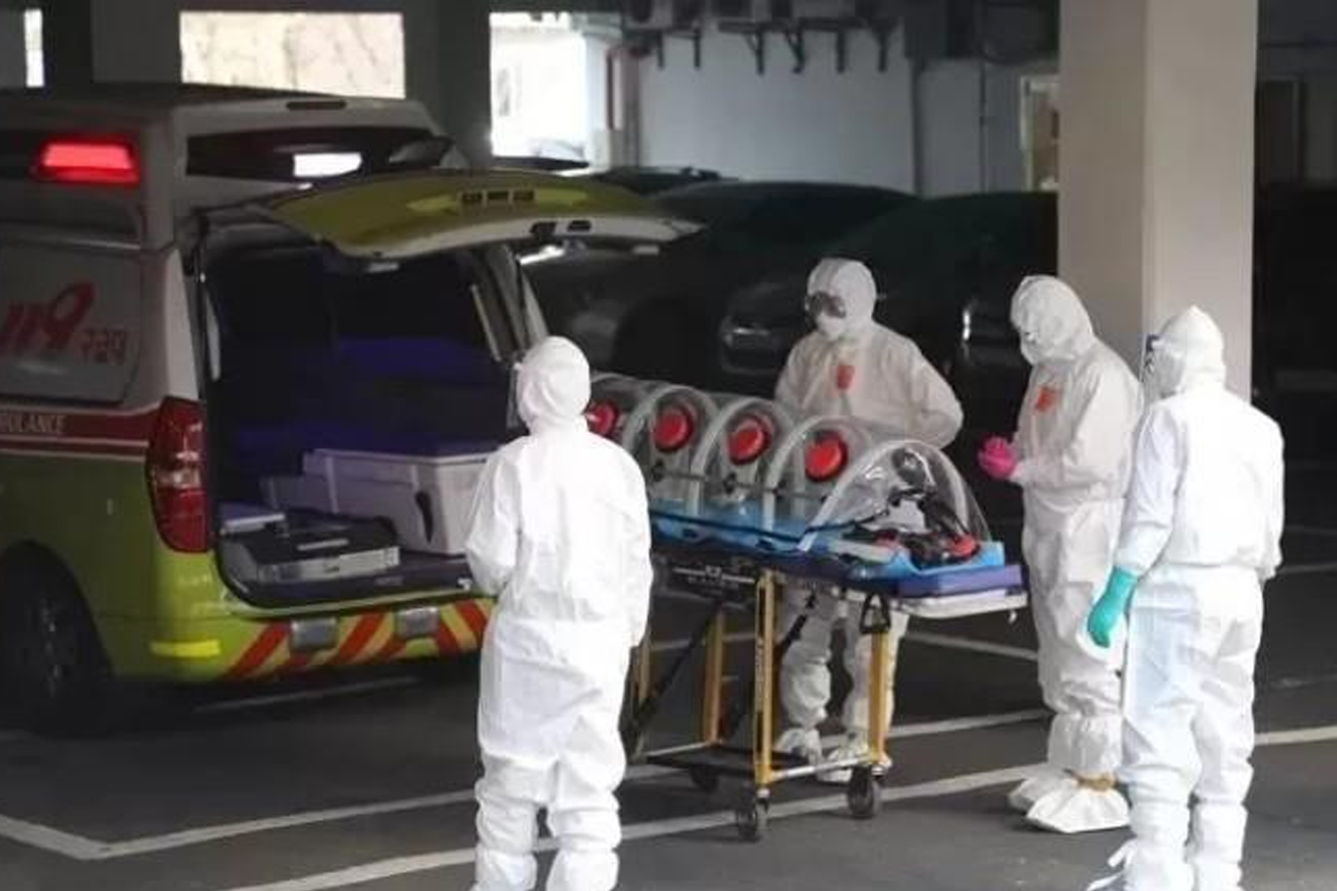 穿著全身防護裝的醫護人員正在推著帶隔離裝置的擔架，背景是停在停車場內的救護車，可能是與COVID-19相關的情景。