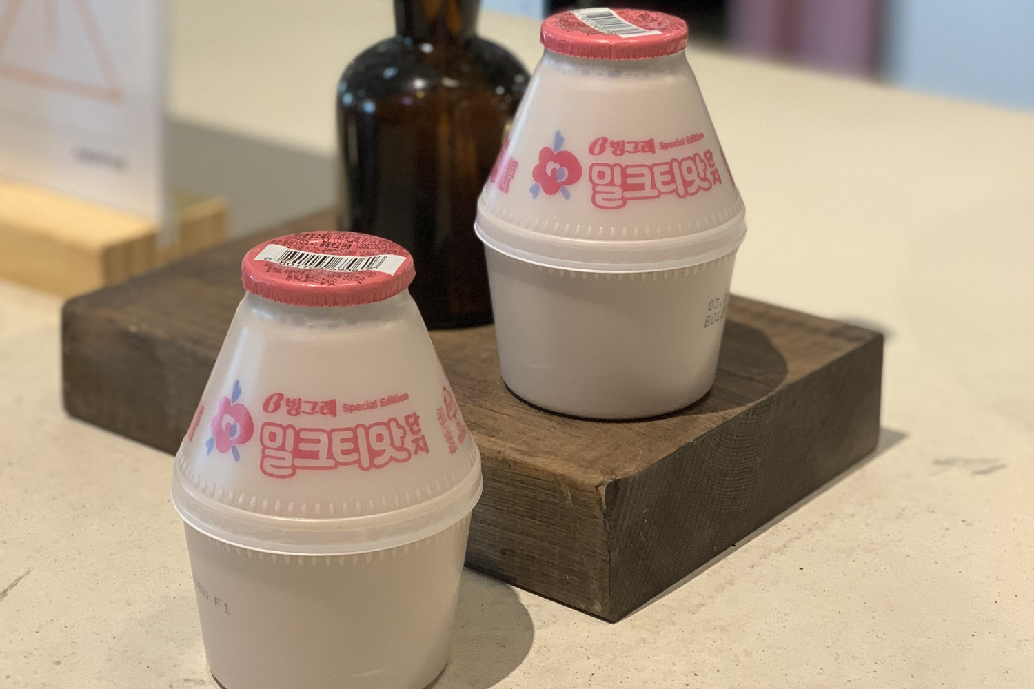 兩瓶裝有特別版韓國香蕉奶的飲品，放在木桌上，旁邊放有深色玻璃瓶，背景是韓式風格的咖啡廳環境。