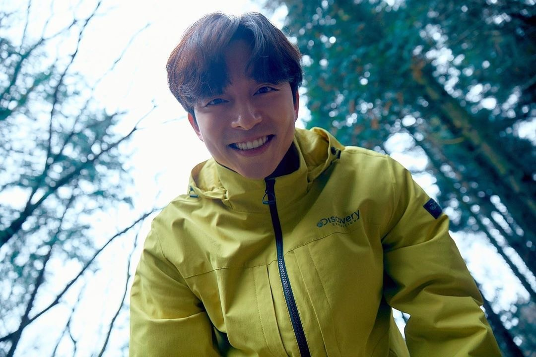 孔刘透过手中相机看向镜头，身穿亮绿色Discovery夹克，与森林背景形成鲜明对比，表现自然探索的主题。