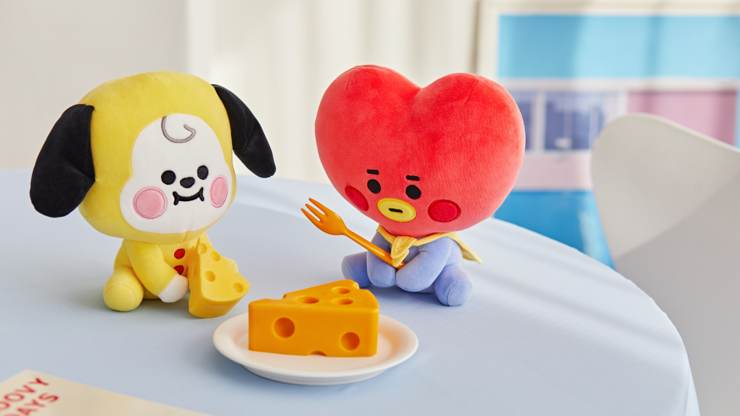 BT21玩偶CHIMMY和TATA坐在桌子旁，桌上摆放着一盘切成块的奶酪。这些可爱的玩偶为任何空间增添了乐趣和互动性。