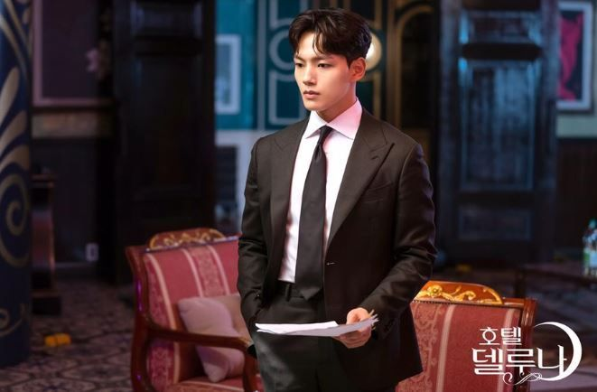 身著西裝的男士站在華麗的酒店背景前，他手持一份文稿，背景展現酒店的典雅裝飾。右下角有德魯納酒店的字樣。