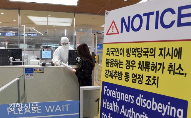仁川国際空港2ターミナルで防護服を着た職員が旅行者を案内する様子。背景に隔離措置に関する注意書があり、入国プロセスの厳格さを強調しています。