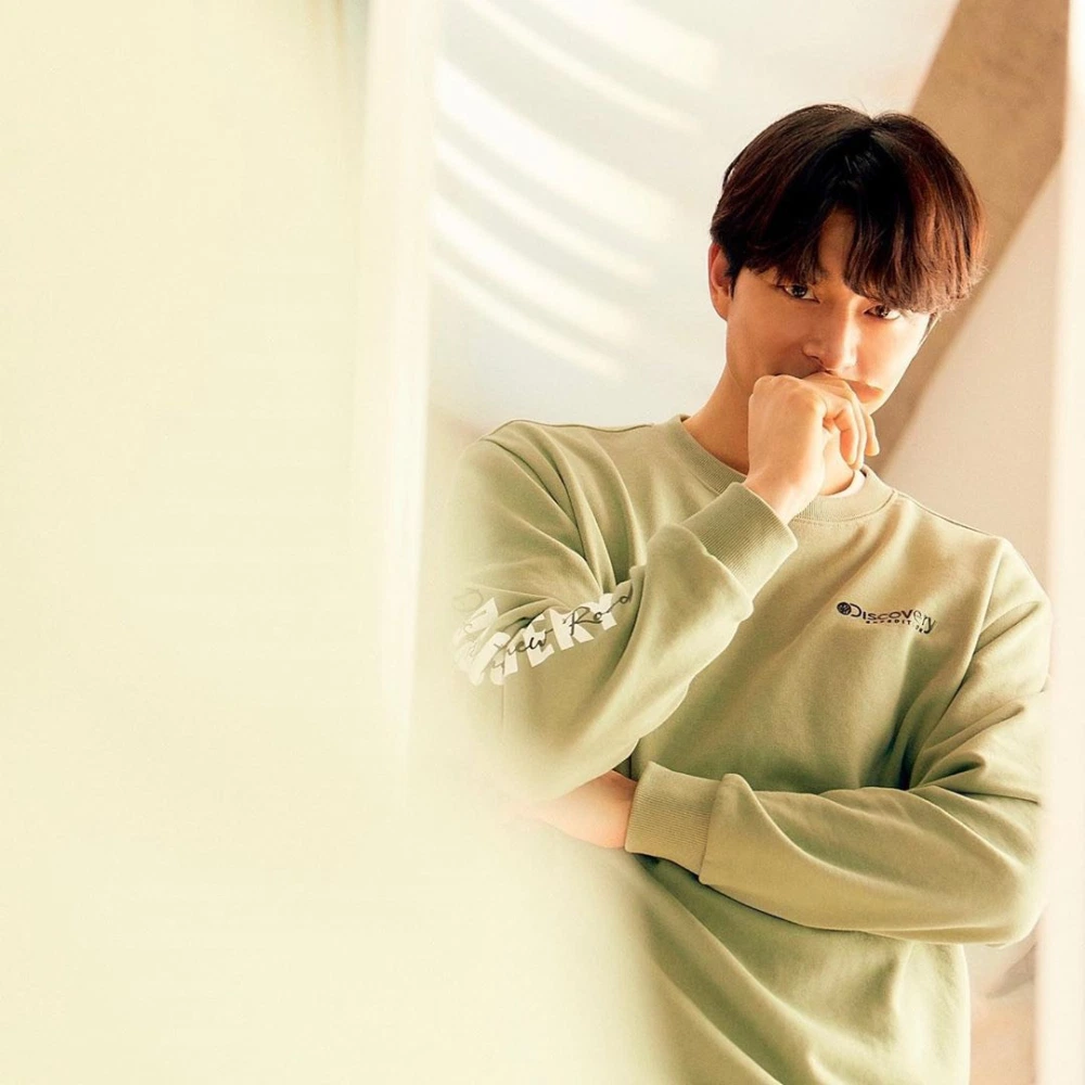 孔刘倚靠桌边，眼神温柔，Discovery品牌蓝色卫衣尽显成熟风采，环境光影交织营造出温馨气氛。