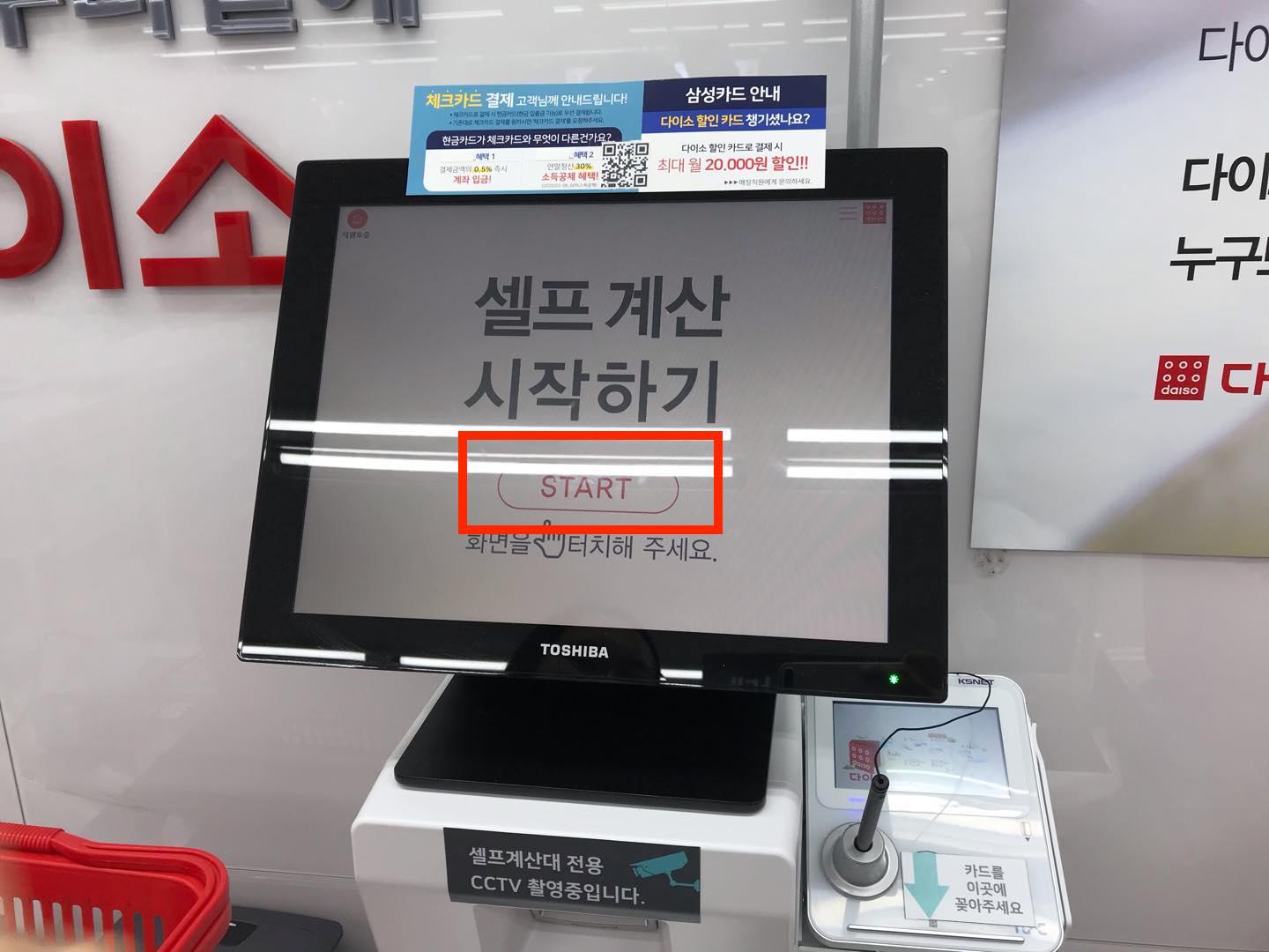 ภาพแสดงเครื่องจ่ายเงินอัตโนมัติที่ร้านไดโซะในเกาหลี โดยมีหน้าจอที่แสดงขั้นตอนแรกของการใช้งานเครื่อง พร้อมกับบาร์โค้ดสแกนเนอร์ที่ใช้ในการตรวจสอบราคาสินค้า