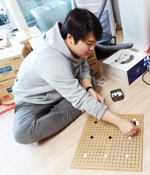 囲碁盤の前に座っている灰色のパーカーを着た人物。周囲に段ボール箱や掃除機が見える室内。