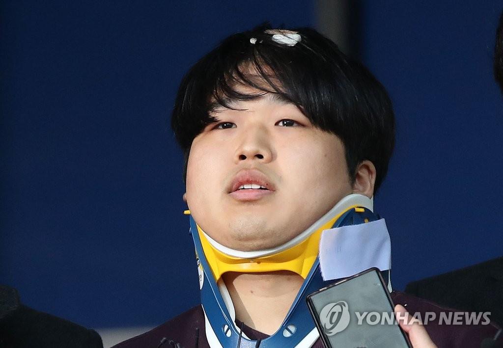 ネックブレースを着用した人物の映像。さまざまな角度から記者たちの録音機が見える。背景は青色の壁が見える。