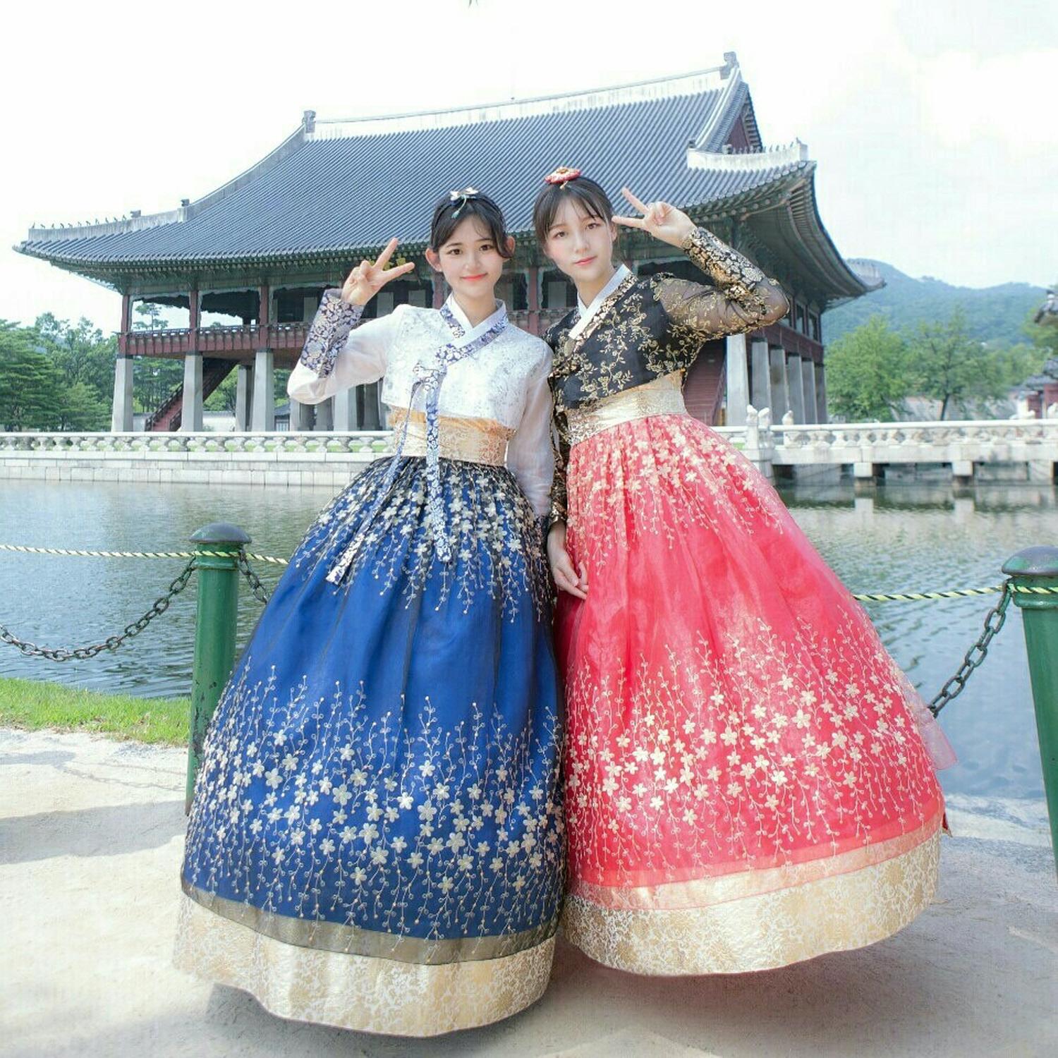 Smiling women in traditional Korean dress stands in front of temple at Gyeongbokgung Palace, with sky and trees in background.