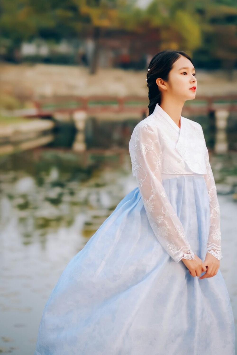 Smiling woman in colorful traditional Korean dress posing in front of a palace with plants and water features.