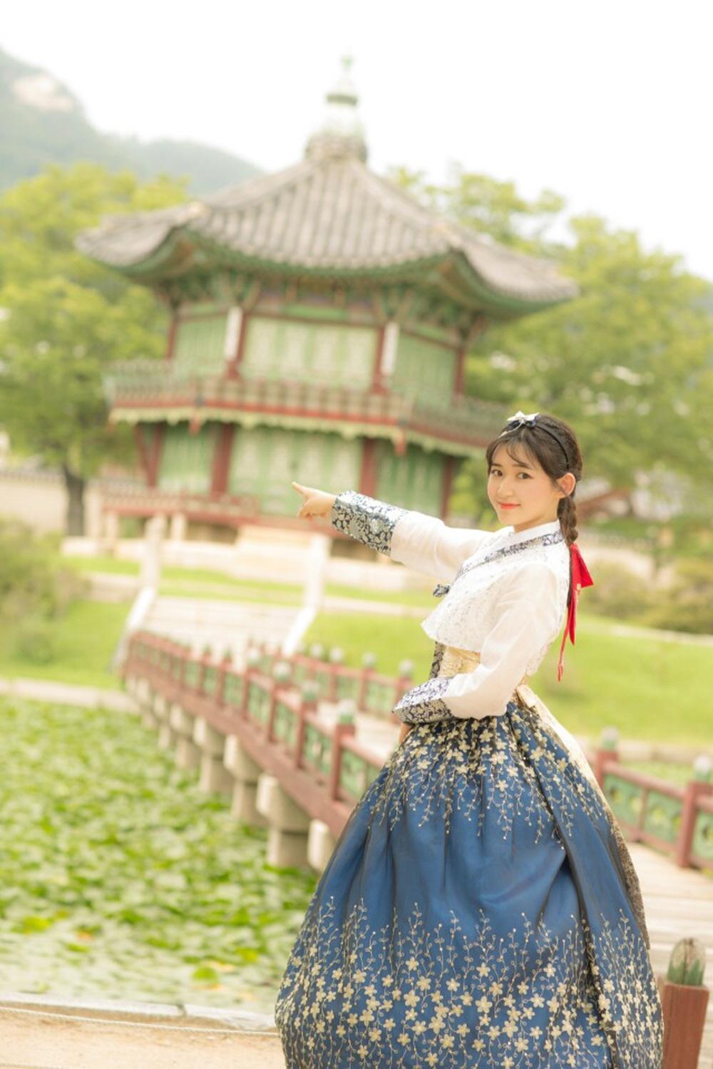 Smiling woman wearing traditional Korean hanbok in front of historic building and trees under blue sky.