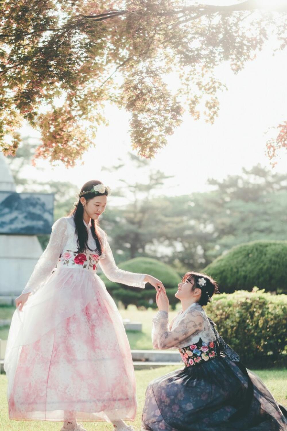 Happy people wearing traditional Korean clothing, posing under trees and plants in front of a palace.