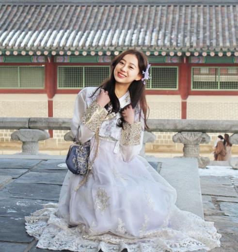 Smiling Korean girl dressed in traditional hanbok with flowing sleeves, standing in front of the beautiful temple gates of Gyeongbokgung Palace in Seoul, Korea.