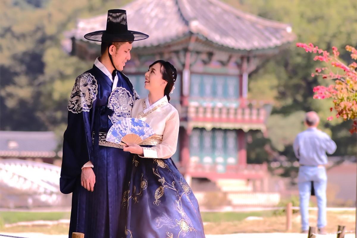 A couple in colorful traditional Korean dresses pose in front of Gyeongbokgung Palace, wearing sun hats and holding a plant and a hat while smiling.