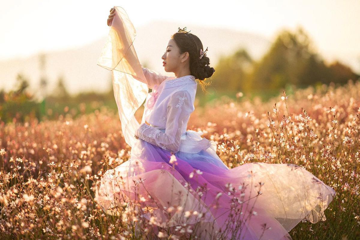 People in hanbok stand in a field of plants and flowers beneath blue sky.