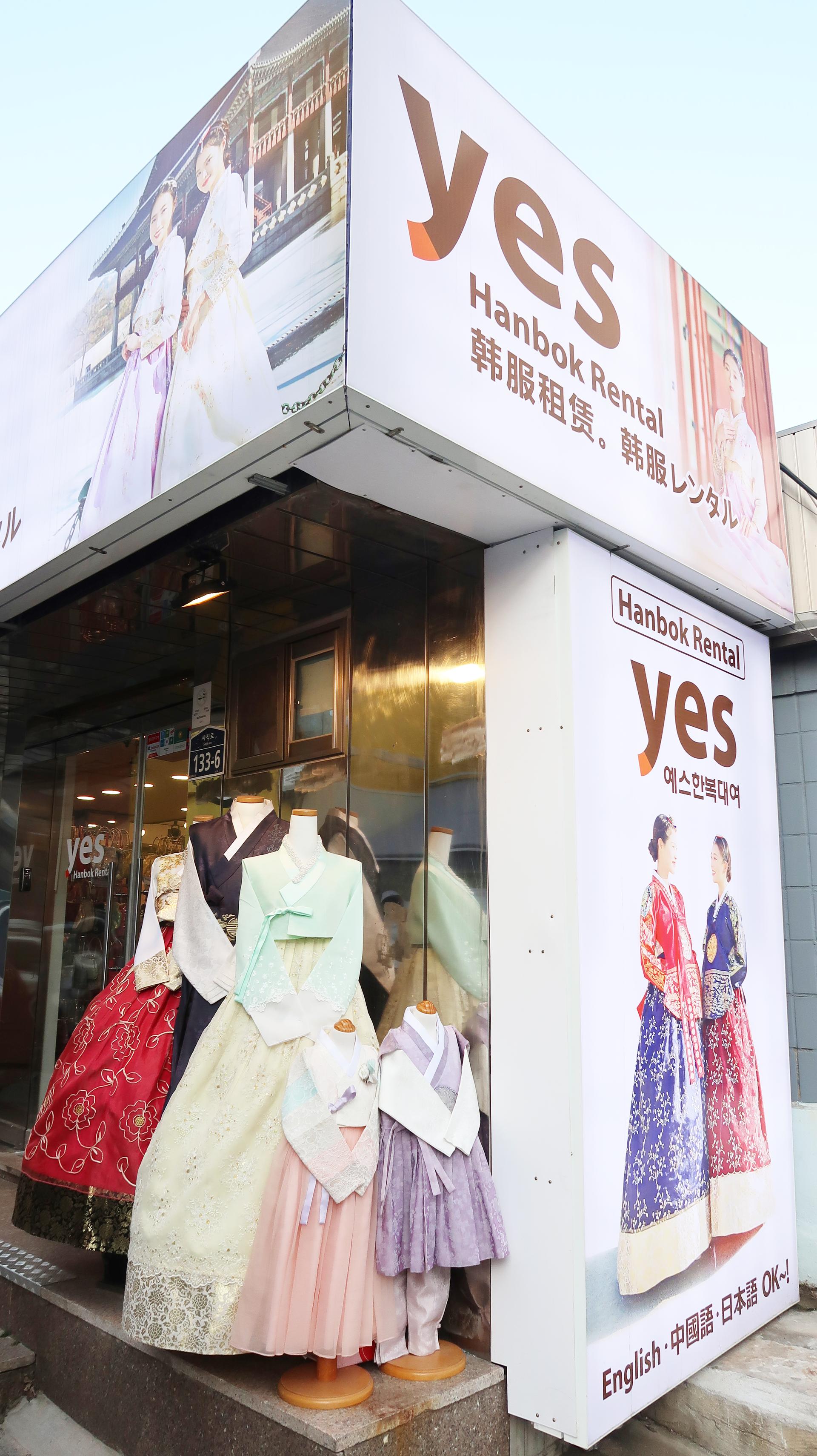 A storefront for 예스한복 in Seoul, offering traditional Korean fashion design, with a bright white facade and a billboard featuring a model wearing their clothing.