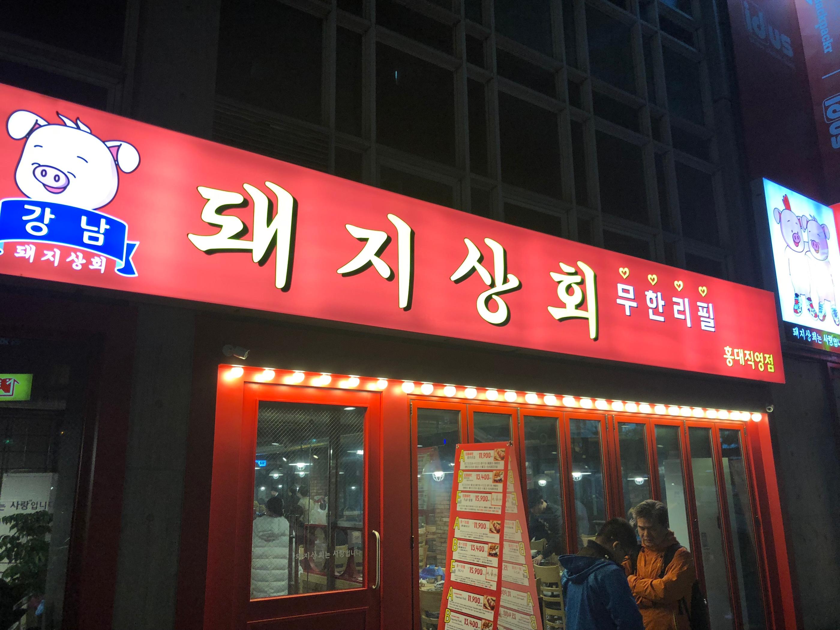 Infinite refill restaurant facade with neon signage and electronic display device at Gangnam Dwaeji Sanghoe in Hongdae, Korea.