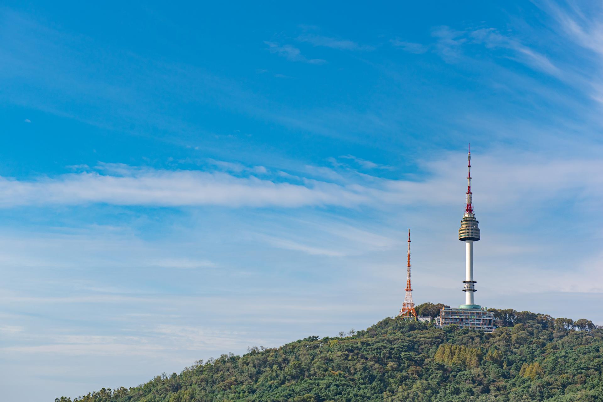 View of N Seoul Tower and its surrounding natural landscape, with clouds and blue sky.