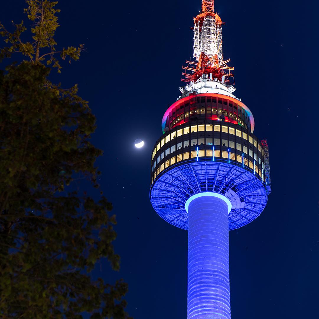 View of N Seoul Tower observation deck at midnight with city skyline, skyscrapers, and moon in background.